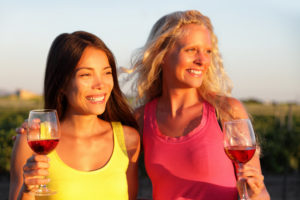 Winery wine tasting tour at countryside vineyard girl friends drinking red wine together watching sunset. Two happy women enjoying summer outdoor activity, multiracial group.